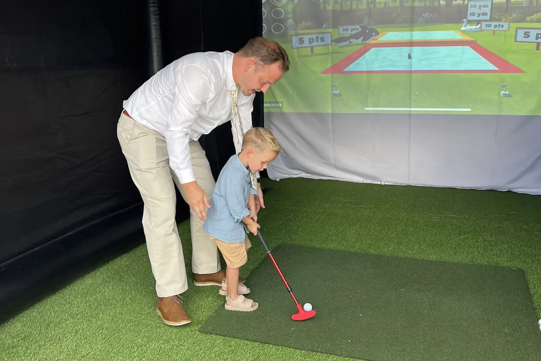 A dad steadying a toddler's putter on the turf inside the Golf to Go Events bay, Shark Splash on the screen behind them