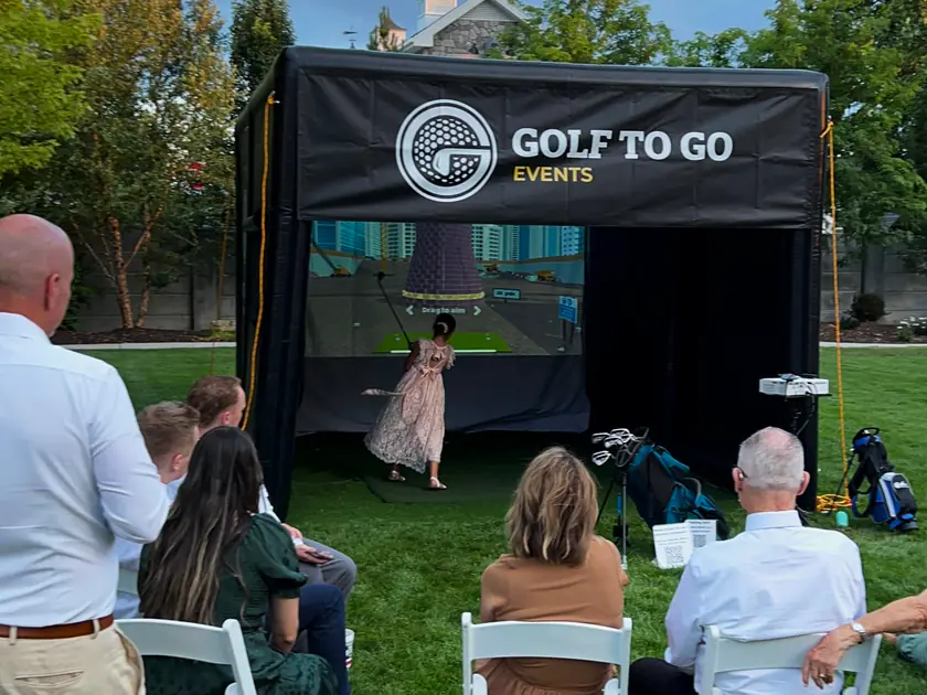 Guests in white chairs on a backyard lawn watching a woman swing inside the Golf to Go Events tent