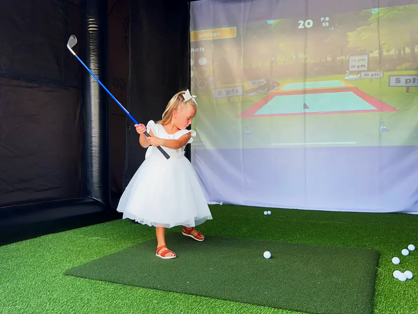 Young guest in a white dress swinging a club on a golf simulator at a Sandy party