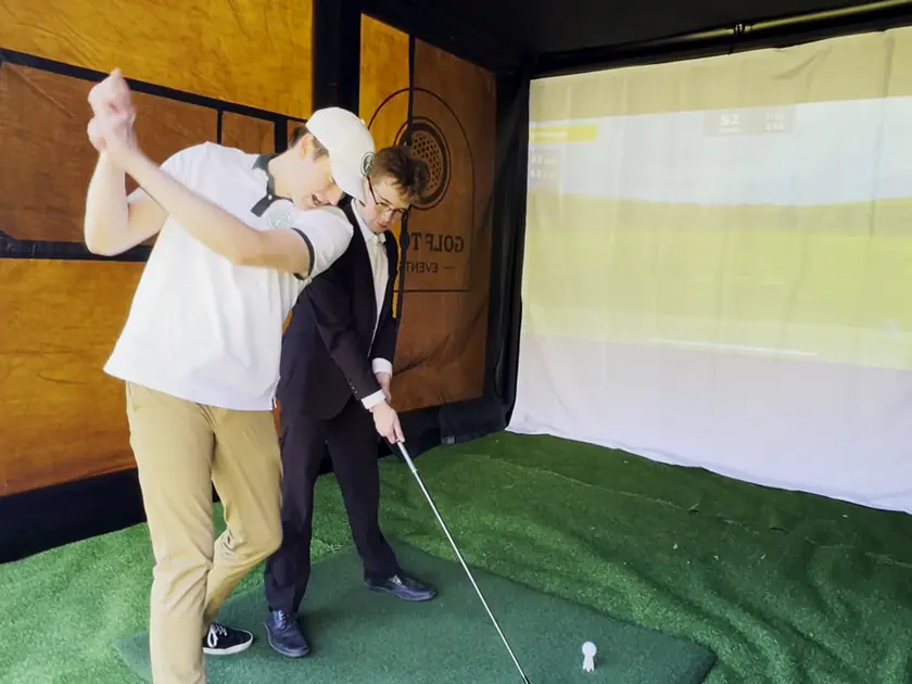 Coworker coaching a colleague through a swing on a mobile golf simulator at a company party