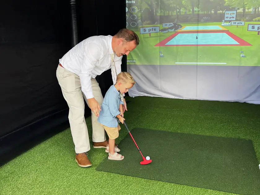 Dad helping his toddler son putt on a portable golf simulator at an Orem, Utah family party